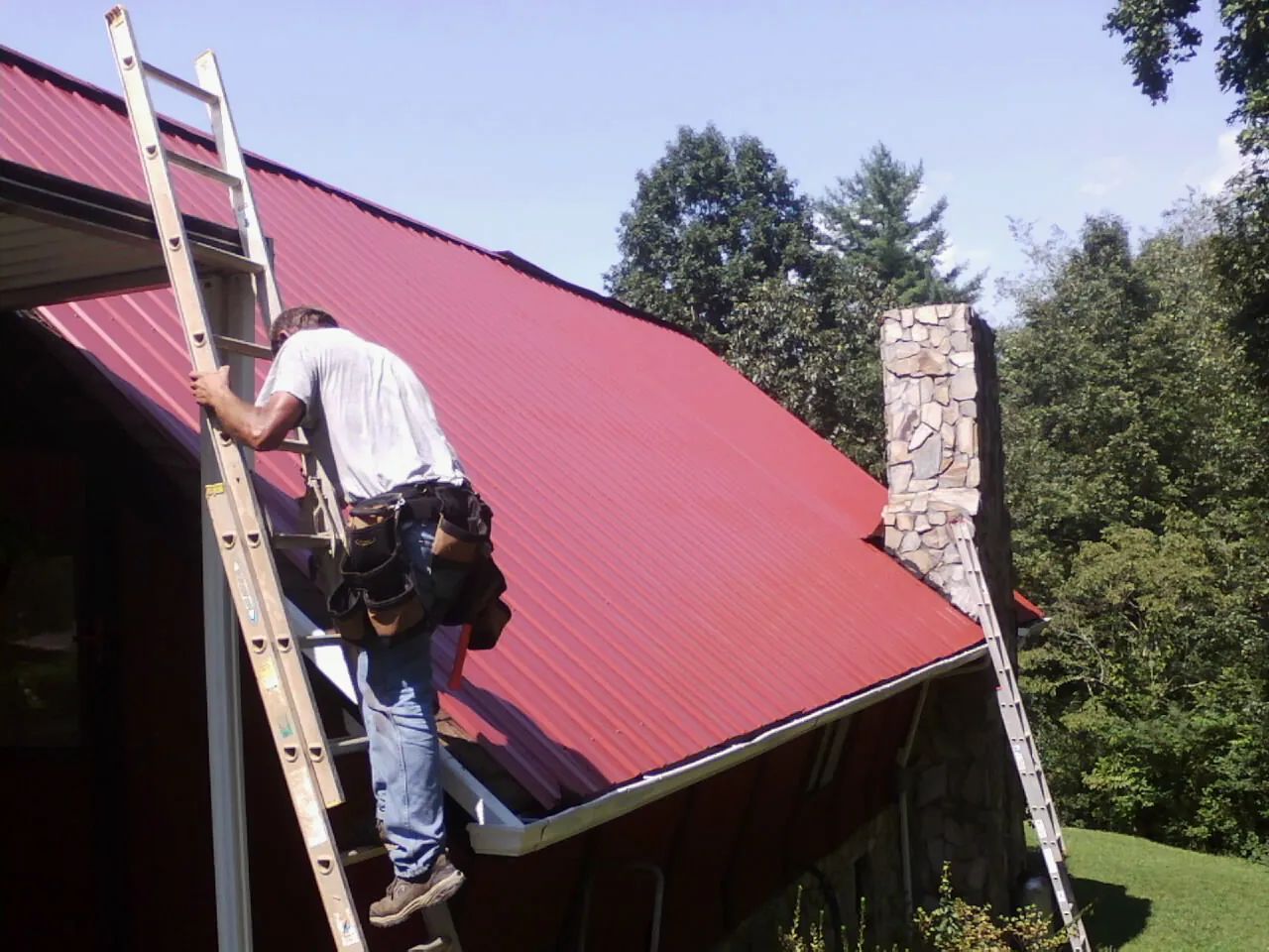 Roofer on a ladder beside a red metal roof and stone chimney — Soffit & Fascia Repair by Roofing Brothers in Mosier
